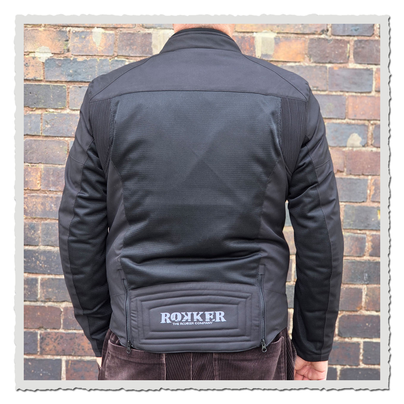 Rückansicht einer Person in schwarzer Jacke vor einer braunen Backsteinwand.